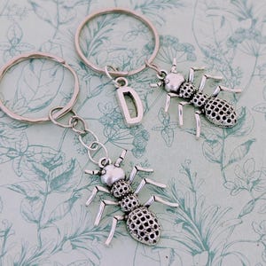 May include: Two silver keychains with ant charms. One keychain has a silver letter D charm. The keychains are on a floral patterned background.