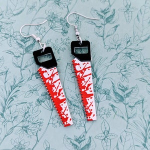 May include: A pair of silver earrings shaped like a bloody saw. The earrings are black with red paint splatters.