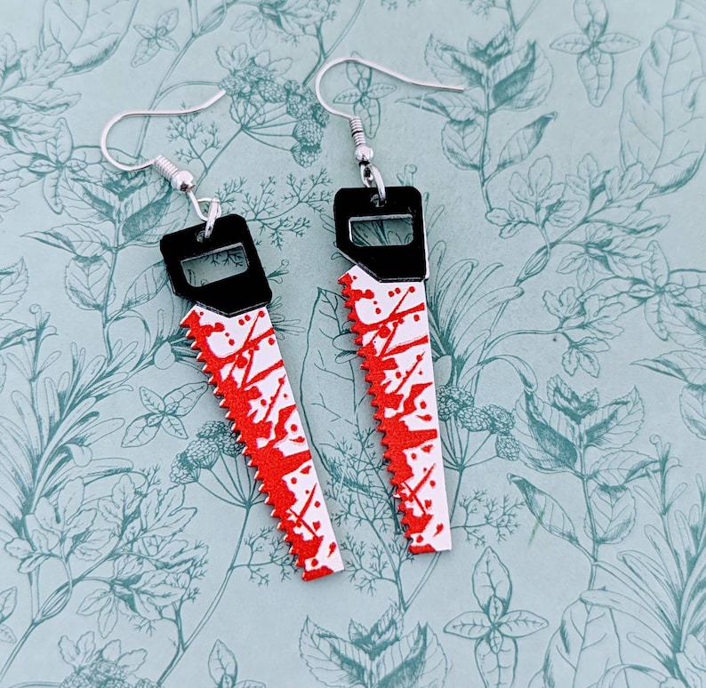 May include: A pair of silver earrings shaped like a bloody saw. The earrings are black with red paint splatters.