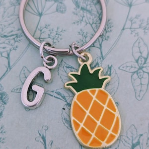 Può includere: Un portachiavi argentato con un ciondolo a forma di ananas e un ciondolo a forma di lettera G argentato. L'ananas è giallo con la parte superiore verde e linee marroni.