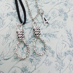 May include: Two silver necklaces with a noose pendant. One necklace has a black cord and the other has a silver chain. The silver chain necklace has a silver charm with the letter A on it.