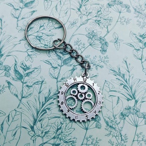 Puede incluir: Llavero plateado de estilo steampunk con un colgante en forma de engranaje con engranajes internos visibles y una esfera de reloj numerada del 1 al 12. El llavero incluye una cadena y un llavero grande. El fondo es un estampado floral azul claro.