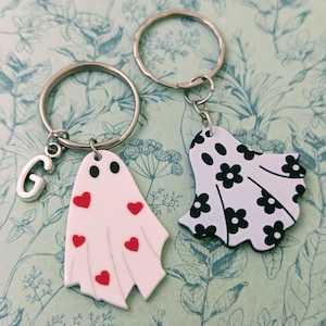 May include: Two ghost-shaped keyrings. One is white with red hearts and the other is white with black floral print. Both have silver key rings.