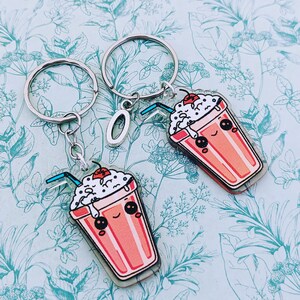 May include: Two silver keychains with a cute cartoon milkshake charm. Each charm has a smiling face, whipped cream, and a straw.