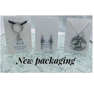 May include: Three silver jewellery pieces displayed on white card stock. The first piece is a silver charm with a sailboat design on a silver key chain. The second piece is a pair of silver earrings with a skeleton design. The third piece is a silver necklace with a round pendant featuring a sailboat design. The card stock has the text "Melspridejewellery" printed on it.