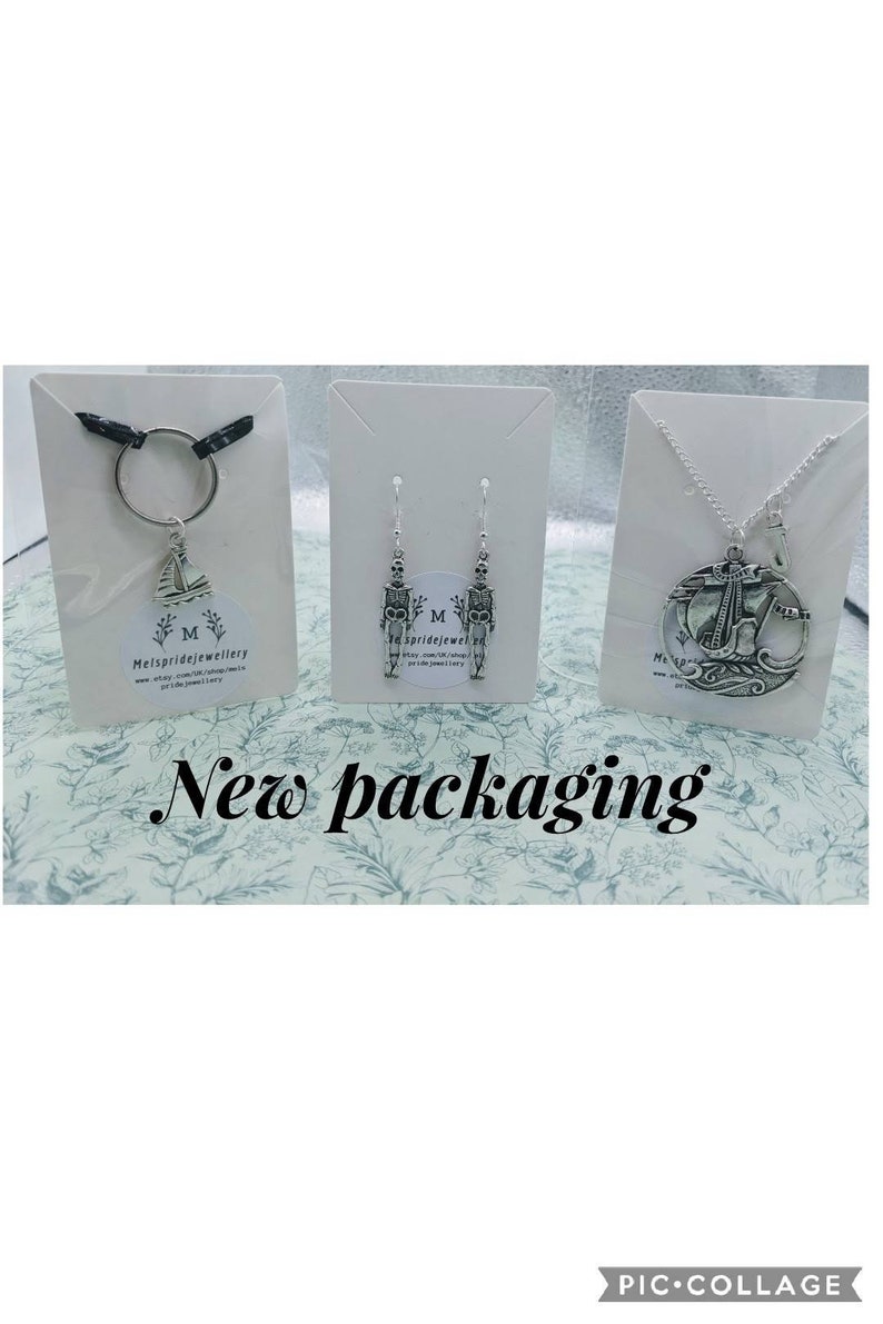 May include: Three silver jewellery pieces displayed on white card stock. The first piece is a silver charm with a sailboat design on a silver key chain. The second piece is a pair of silver earrings with a skeleton design. The third piece is a silver necklace with a round pendant featuring a sailboat design. The card stock has the text "Melspridejewellery" printed on it.