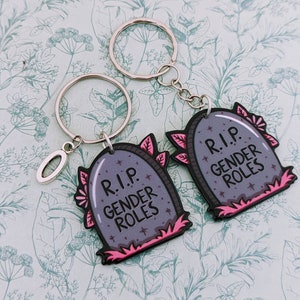 May include: Two silver keychains with black and pink tombstone-shaped charms. The charms have the text "R.I.P. Gender Roles" on them.