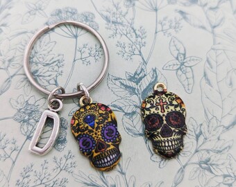 May include: A silver key chain with a letter D charm and two colorful sugar skull charms. The sugar skulls are decorated with intricate designs and have black eyes and a cross on the forehead.