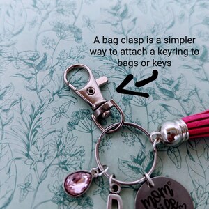May include: A silver keyring with a pink crystal charm, a silver letter D charm, and a silver charm with the text "mom wife boss" on it. The keyring is attached to a red tassel.