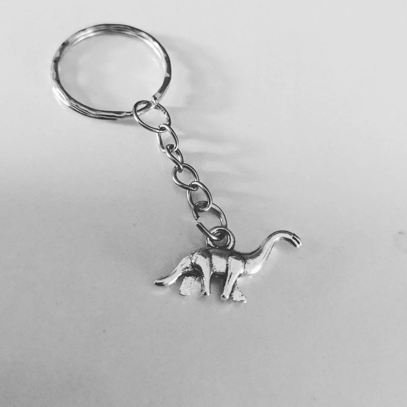 May include: Silver keychain featuring a dinosaur charm. The dinosaur is standing on all fours with its neck raised and head tilted slightly to the side.