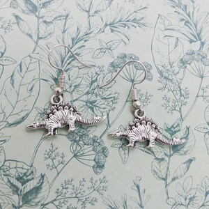 May include: A pair of silver dangle earrings featuring a dinosaur charm. The dinosaur is a stegosaurus with a spiky back and a long tail.