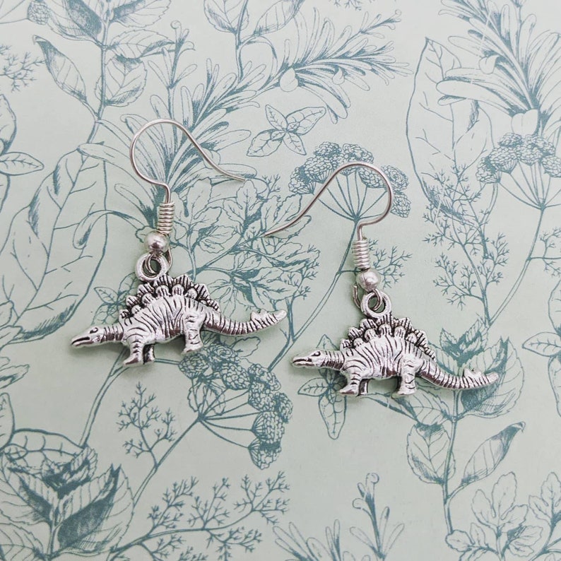 May include: A pair of silver dangle earrings featuring a dinosaur charm. The dinosaur is a stegosaurus with a spiky back and a long tail.