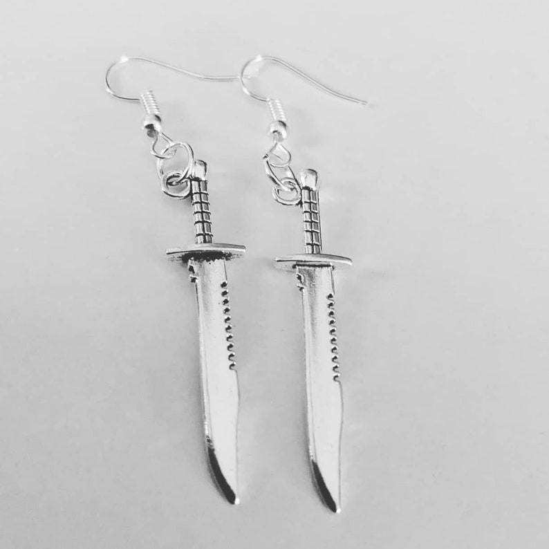 May include: A pair of silver earrings shaped like swords. The swords have a serrated edge and a pointed tip.