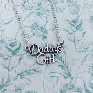 May include: Silver-toned necklace with the words "Daddy's Girl" in a cursive font. The pendant hangs from a delicate chain. The background features a light blue floral pattern.