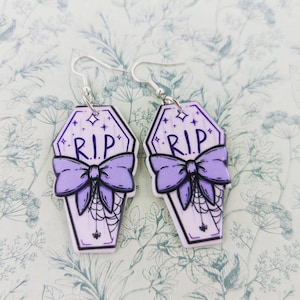 May include: A pair of purple coffin-shaped earrings with a black bow and the text "RIP" on each earring. The earrings have silver hooks.