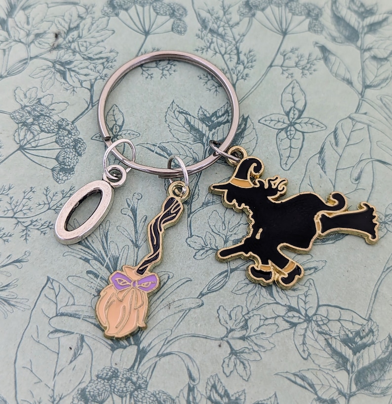 May include: A silver key chain with a black witch charm, a gold broom charm, and a silver charm shaped like the letter "O".