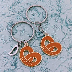 Op de afbeelding: Twee zilveren sleutelhangers met een pretzel-bedel. Een sleutelhanger heeft een letter D-bedel. De bedels zijn oranje en bruin.