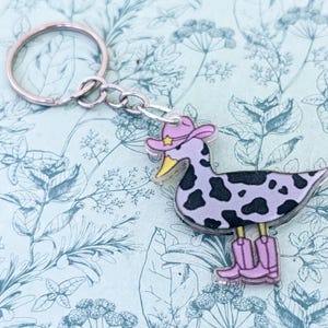 Puede incluir: Un llavero novedoso con un diseño de pato con sombrero y botas de vaquero. El pato tiene un cuerpo lavanda con manchas negras, un pico amarillo y está unido a un llavero plateado.