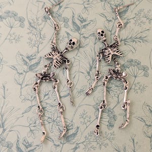 Puede incluir: Un par de pendientes de esqueleto plateados. Cada pendiente presenta un diseño de esqueleto detallado, que incluye un cráneo, una caja torácica y extremidades articuladas. Los pendientes cuelgan de un gancho simple. El fondo es un patrón floral verde claro.
