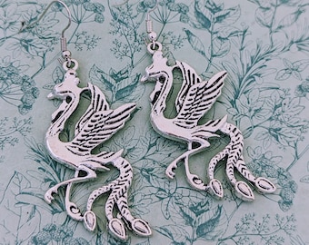 May include: A pair of silver earrings featuring a detailed design of a phoenix with outstretched wings and a long tail.