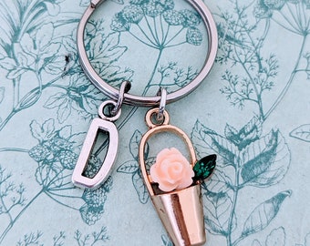 May include: A silver key ring with a charm of a small gold bucket with a pink rose and green leaves. The letter "D" is also attached to the key ring.