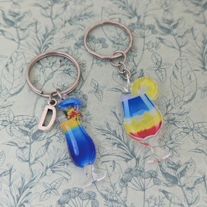 May include: Two silver keychains with colorful glass charms shaped like cocktail glasses. One charm has a blue and yellow drink with a flower garnish and the letter "D" on the keychain. The other charm has a red, yellow, and blue drink with a lemon garnish.