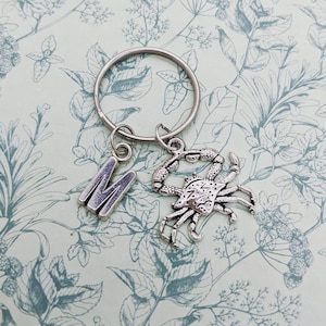 May include: Silver keychain with a letter M charm and a crab charm. The charms are attached to a silver key ring.