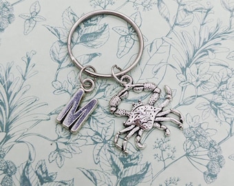May include: Silver keychain with a letter M charm and a crab charm. The charms are attached to a silver key ring.