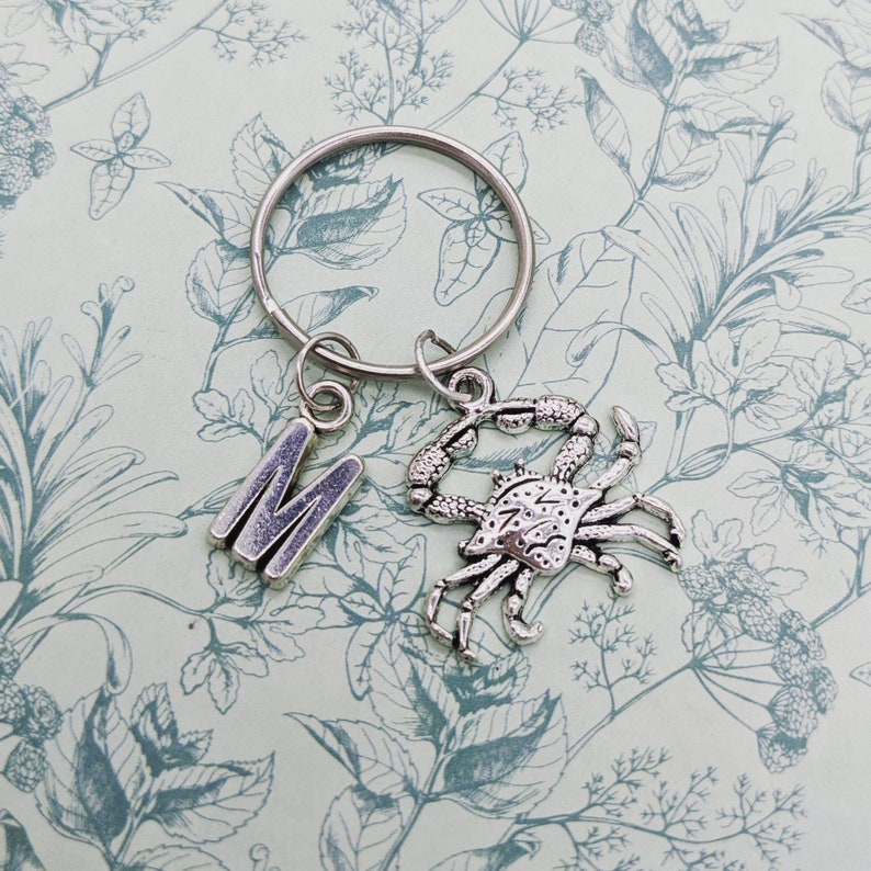 May include: Silver keychain with a letter M charm and a crab charm. The charms are attached to a silver key ring.