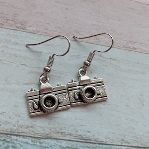 May include: A pair of silver camera earrings. The earrings are shaped like a vintage camera with a round lens and a silver hook.