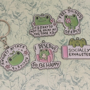 May include: A collection of green frog-themed charms and a keychain. The charms feature phrases like "Anxiety Attack Survivor," "I Deserve to Be Happy," and "Socially Exhausted." Each charm has a unique design with a cute frog illustration.