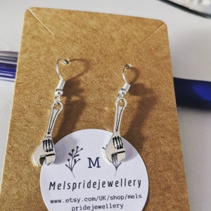 May include: Silver adjustable wrench earrings on a brown card with the text "M" and "Melspridejewellery" and the website address "www.etsy.com/UK/shop/mels pridejewellery".