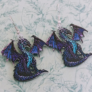 Pode incluir: Um par de brincos pendentes com um design de dragão roxo e azul. Os dragões têm asas iridescentes e estão empoleirados em um gancho prateado.