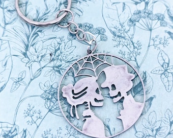 May include: Silver skeleton couple keychain with a spiderweb design. The skeletons are facing each other and holding hands. The keychain is attached to a silver ring.
