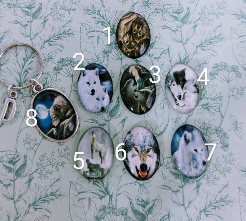 May include: Eight round glass charms featuring images of wolves, each charm has a number from 1 to 8 printed on the background. The charms are displayed on a white background with green leafy patterns.