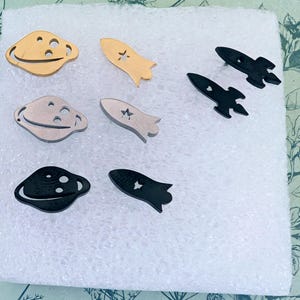 Puede incluir: Una colección de dijes de metal con temática espacial que incluye tres formas de planetas y cinco formas de cohetes. Los dijes tienen acabados en tonos oro, plata y negro, cada uno con detalles recortados como pequeños círculos y estrellas.