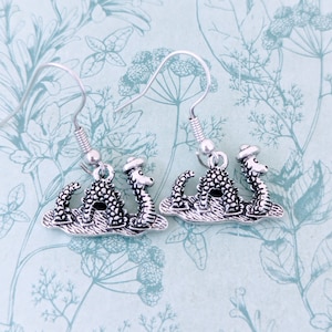 May include: Silver dangle earrings featuring a small, detailed, three-dimensional sculpture of a mythical sea serpent wearing a hat.
