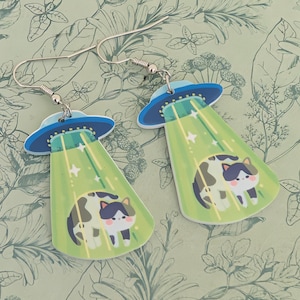 Peut inclure: Boucles d'oreilles fantaisie représentant un chat de dessin animé enlevé par un OVNI. Les boucles d'oreilles sont bleues et vertes, avec un crochet argenté. Le chat est noir, blanc et marron.