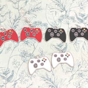 以下が含まれることがあります： 赤、黒、白のミニチュアゲームコントローラーのイヤリング。各コントローラーにはシルバーのフックと詳細なボタンデザインがあります。イヤリングは花柄の背景に配置されています。