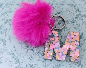 May include: A pink fluffy keychain with a silver key ring attached to a clear resin letter M filled with iridescent pink, gold, and white glitter hearts.
