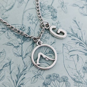 May include: A silver necklace with a charm of a person doing a downward-facing dog yoga pose and a charm of the letter G.