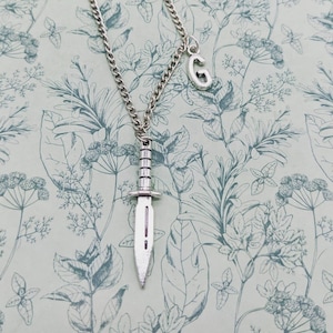 Peut inclure: Un collier en argent avec un pendentif en forme de couteau et un charme en forme de lettre 'G'. Le collier est sur un fond à motifs floraux.