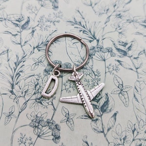 Puede incluir: Llavero de avión plateado con un dije de letra D plateada. El llavero está sobre un fondo con estampado floral.