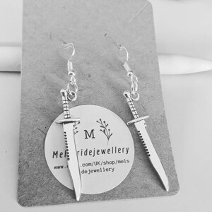 May include: A pair of silver earrings featuring a sword design. The swords have a serrated edge and are hanging from silver hooks.