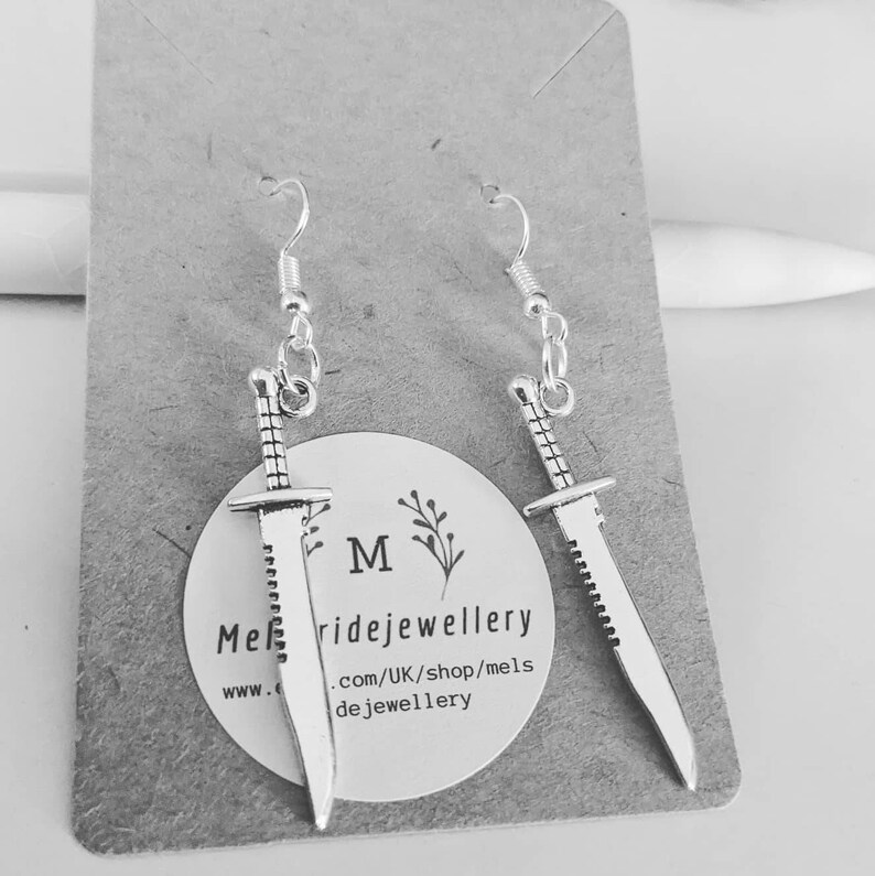 May include: A pair of silver earrings featuring a sword design. The swords have a serrated edge and are hanging from silver hooks.