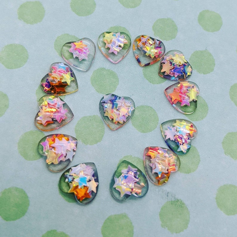 May include: A set of 12 iridescent heart-shaped charms with a rainbow star design. The charms are made of clear plastic and have a holographic finish.