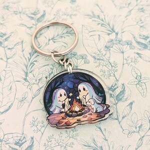 May include: A round keychain featuring a whimsical illustration of two ghosts and a dog gathered around a campfire under a starry night sky. The keychain has a silver metal ring and clasp. The background is a blue and purple night scene.