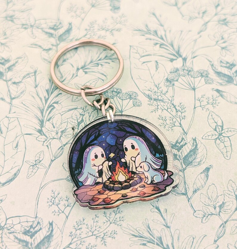 May include: A round keychain featuring a whimsical illustration of two ghosts and a dog gathered around a campfire under a starry night sky. The keychain has a silver metal ring and clasp. The background is a blue and purple night scene.
