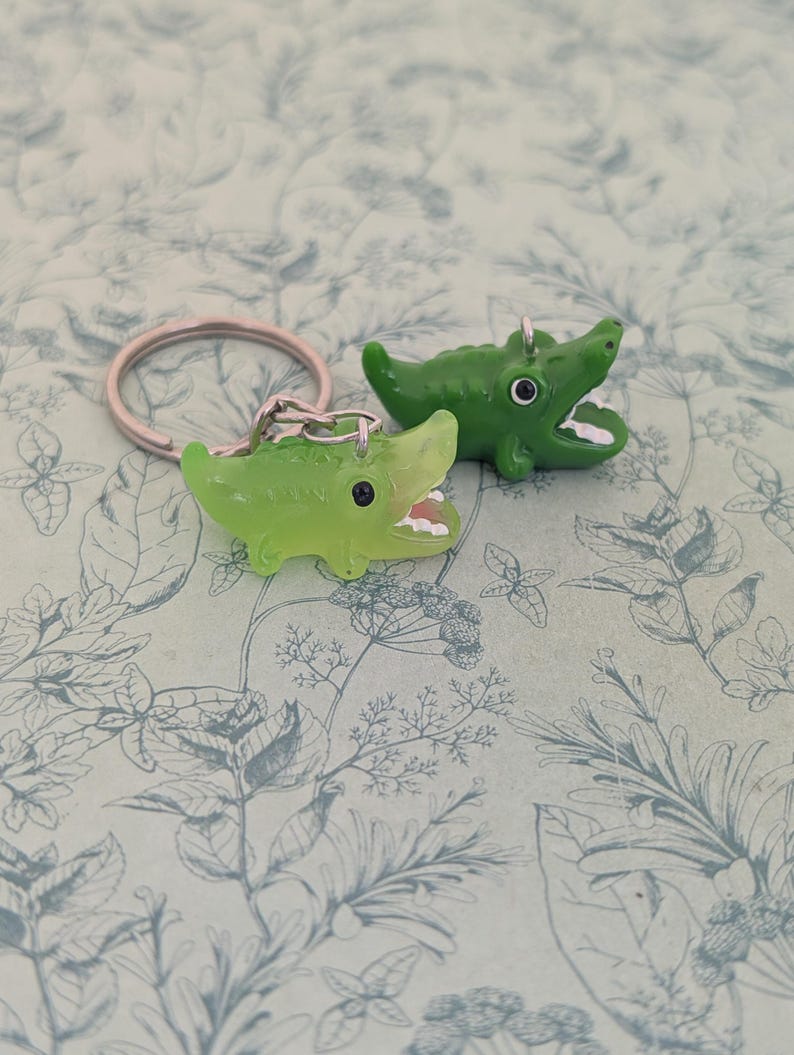 May include: Two green alligator keychains. One keychain is attached to a silver ring. The alligators have open mouths with white teeth. The keychains are on a light blue background with a floral pattern.