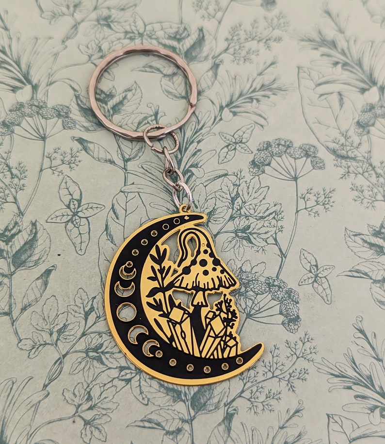 May include: Gold-tone metal keychain featuring a crescent moon design with black enamel. The moon has a mushroom and crystals inside, with a black outline.